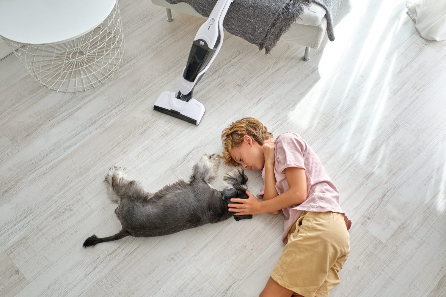 Een kind ligt op de vloer naast een kleine grijze hond, terwijl ze elkaar rustig aanraken in een helder verlichte kamer met een stofzuiger op de achtergrond.